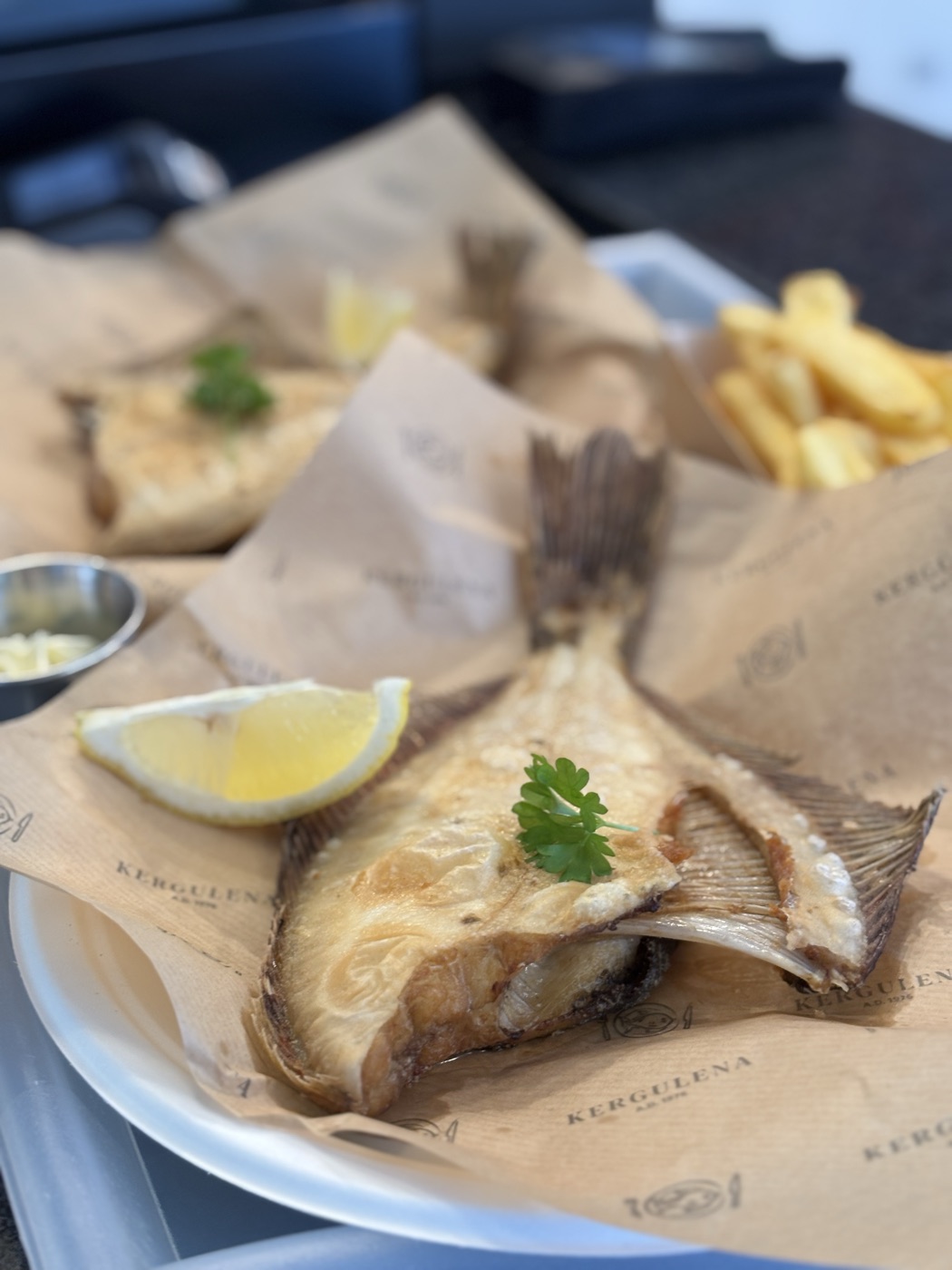 Flądra smażona tradycyjnie — cała ryba z głową i ogonem, z cytryną i natką pietruszki, na papierze Kergulena, z frytkami w tle