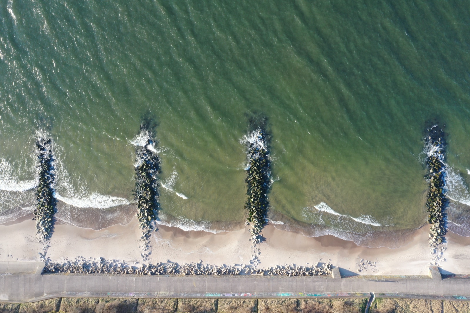 Trzy drewniane falochrony z lotu ptaka — turkusowa woda i piana morska